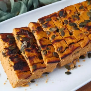 A close-up image of sliced Keto Cottage Cheese Pumpkin Bread topped with pumpkin seeds, presented on a white plate. The bread has a golden-brown crust and a soft, orange-hued interior indicative of pumpkin.