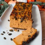 A freshly baked loaf of Keto Cottage Cheese Pumpkin Bread is presented on a white rectangular plate. The bread has a rich, golden-orange hue indicative of pumpkin flavor, with a moist texture visible from the sliced end. It’s topped with scattered pumpkin seeds adding a contrasting green color and texture. The loaf is partially sliced, revealing its dense yet airy crumb structure. In the background, there are subtle hints of autumn decor, suggesting a seasonal theme.