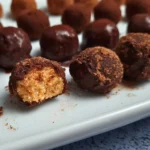A close-up image of Keto Cottage Cheese Pumpkin Balls coated in a dark powder, displayed on a white plate with one ball cut in half to show the creamy orange filling.