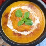 A vibrant orange Keto Pumpkin Soup garnished with a swirl of cream and a sprinkle of spices, served in a black bowl.