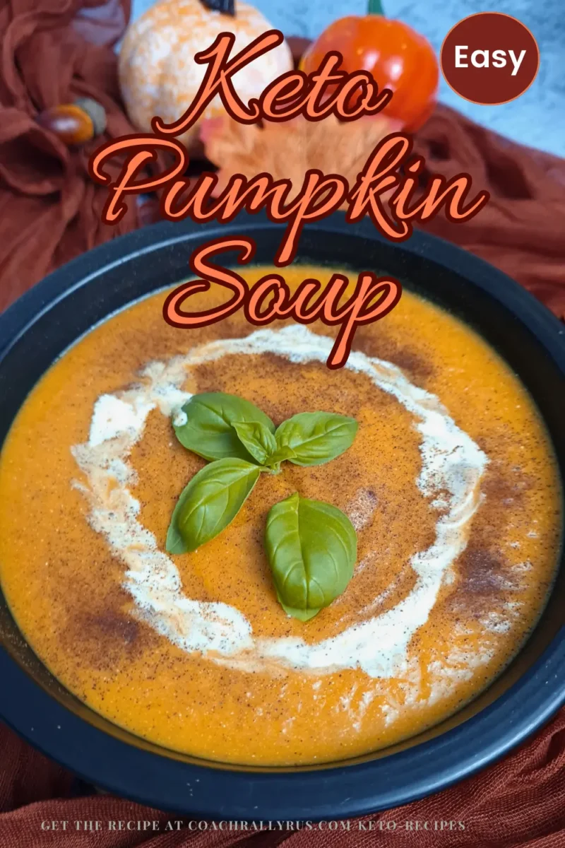 A vibrant orange Keto Pumpkin Soup garnished with a swirl of cream and fresh basil leaves, served in a black bowl against a backdrop of an orange pumpkin and red fabric.