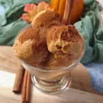 A glass bowl filled with scoops of pumpkin cottage cheese keto ice cream, sprinkled with cinnamon, presented on a wooden surface with autumn leaves and a small pumpkin in the background, illustrating a high-protein, low-carb dessert option.