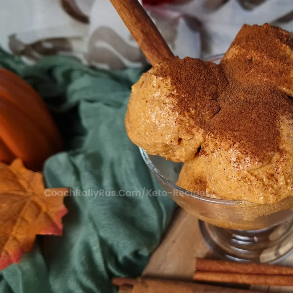 A close-up image of a serving of pumpkin cottage cheese keto ice cream topped with a sprinkle of cinnamon, presented in a clear glass dessert bowl with a wooden spoon. In the background, there is a whole pumpkin, autumn leaves, and cinnamon sticks on a fabric surface, suggesting an autumnal theme.