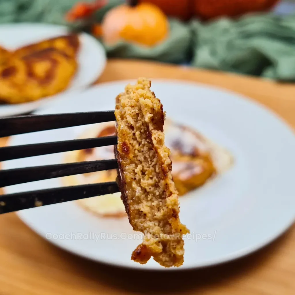 A close-up of a fork holding a bite-sized piece of Keto Pumpkin Spice Cottage Cheese Protein Pancake with more pancakes and pumpkins blurred in the background.