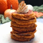 A stack of six golden-brown pumpkin spice cottage cheese protein pancakes on a white plate, garnished with a dollop of cream and a sprinkle of spice on top.