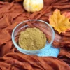 A clear glass bowl filled with pumpkin spice blend sits on a blue cloth, surrounded by an orange-brown fabric that evokes the warmth of autumn. A small white and orange pumpkin and a yellow maple leaf are also visible, adding to the fall theme.