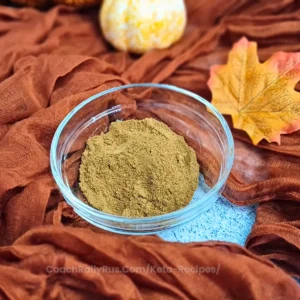 A clear glass bowl filled with pumpkin spice blend sits on a blue cloth, surrounded by an orange-brown fabric that evokes the warmth of autumn. A small white and orange pumpkin and a yellow maple leaf are also visible, adding to the fall theme.