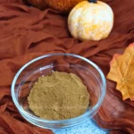 A clear glass bowl filled with pumpkin spice blend sits on a blue cloth, surrounded by an orange-brown fabric that evokes the warmth of autumn. A small white and orange pumpkin and a yellow maple leaf are also visible, adding to the fall theme.