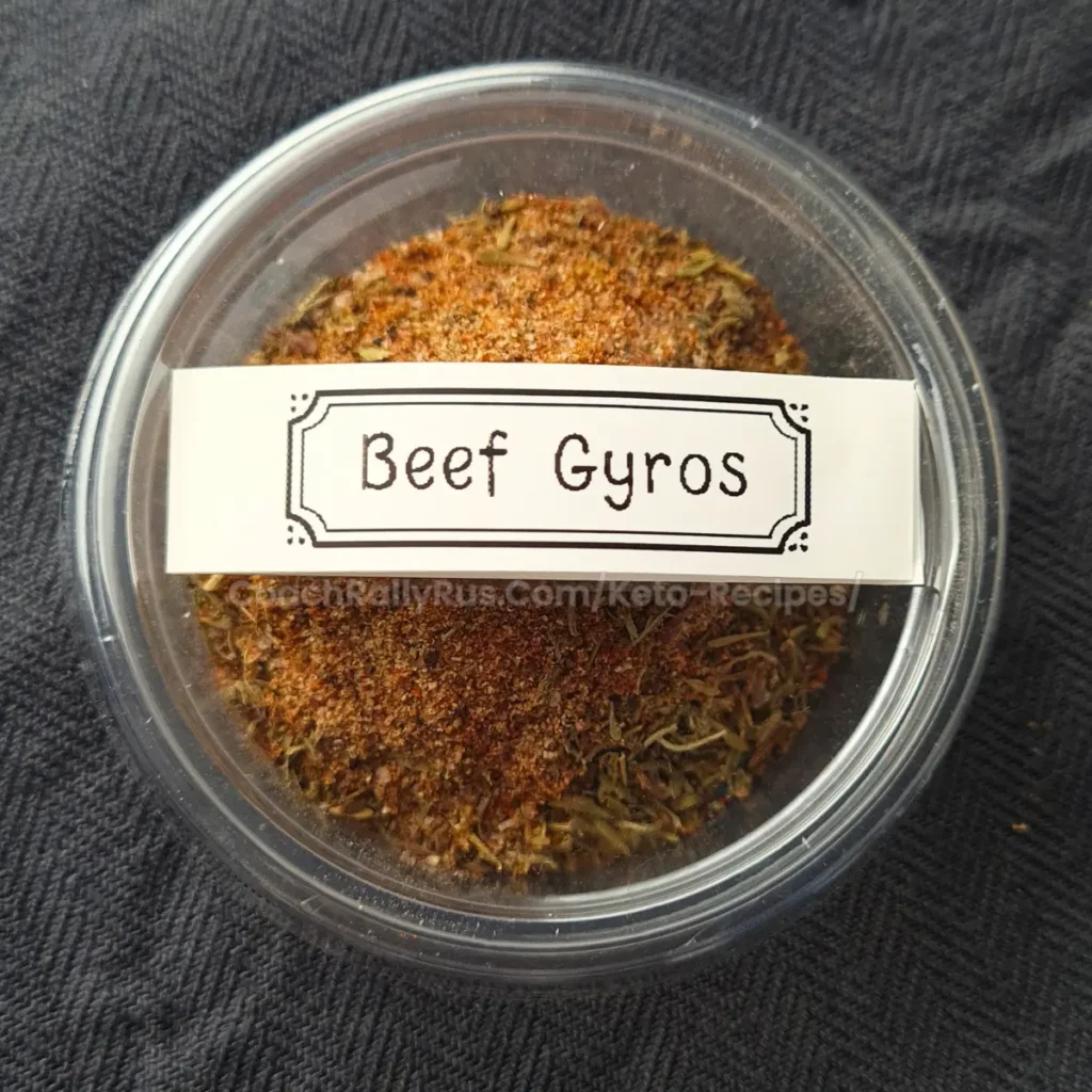 A clear plastic container filled with a mixture of spices labeled ‘Beef Gyros’ on a white label with decorative borders. The container is on a dark fabric surface.