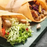 An image showcasing two types of stuffed meat wraps on a wooden surface. On the left is a Gyro with sliced meat, tomatoes, onions, and shredded lettuce wrapped in a thin flatbread. On the right is a Doner Kebab with chunks of meat, purple cabbage, onions, and greens in a thicker bread cone. Both are garnished with fresh vegetables around them.