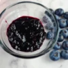 A glass jar filled with dark purple Keto blueberry jam on a white surface, with several whole blueberries scattered around it.