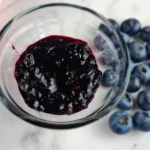 A glass jar filled with dark purple Keto blueberry jam on a white surface, with several whole blueberries scattered around it.