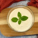 A glass bowl filled with creamy Keto Cottage Cheese Alfredo Sauce, garnished with fresh basil leaves on top, placed on a wooden cutting board.