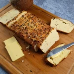 A freshly baked loaf of Keto Cottage Cheese Bread with Coconut Flour, sliced and placed on a wooden cutting board. The bread is topped with a sprinkle of seeds and herbs, giving it a rustic appearance. Beside the bread is a piece with butter on top, a block of butter, and a knife with butter residue.