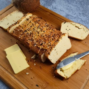A freshly baked loaf of Keto Cottage Cheese Bread with Coconut Flour, sliced and placed on a wooden cutting board. The bread is topped with a sprinkle of seeds and herbs, giving it a rustic appearance. Beside the bread is a piece with butter on top, a block of butter, and a knife with butter residue.