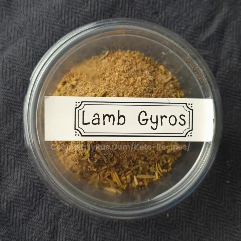 A clear plastic container filled with a mixture of spices labeled “Lamb Gyros” on a black textured background.
