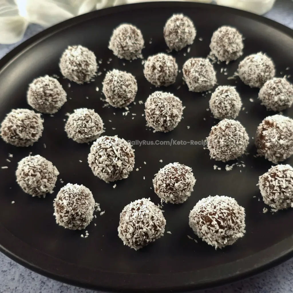 Multiple keto chocolate rum balls on a black plate, rolled in coconut.