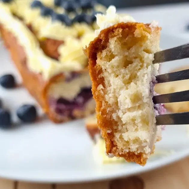 A piece of keto blueberry creme fraiche pound cake with keto Royal Icing Buttercream, garnished with fresh blueberries, being lifted on a fork.