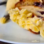 A slice of cake with layers of purple filling and yellow keto royal icing buttercream frosting, next to a piping bag on a white plate.