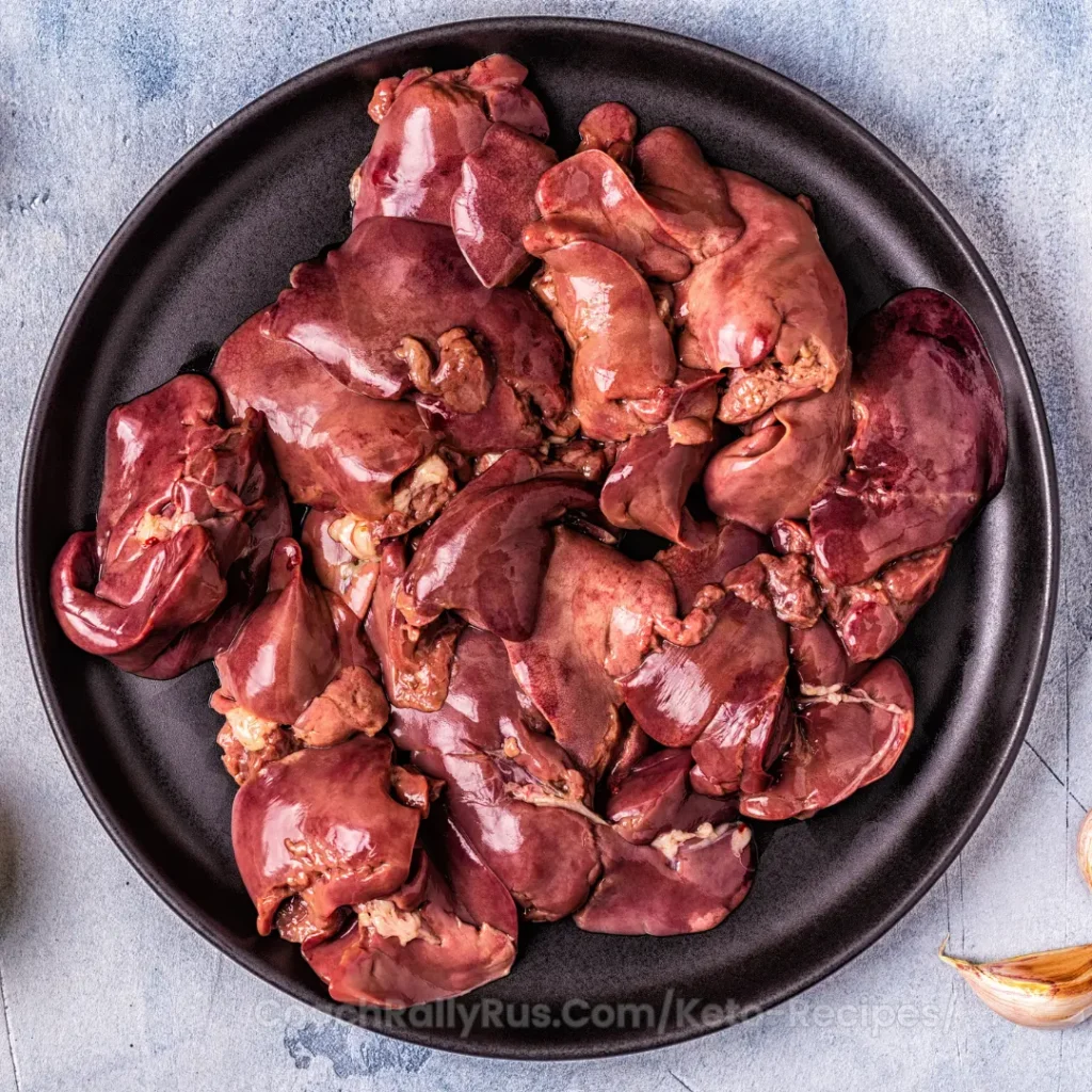 A plate of raw chicken liver pieces on a dark plate with garlic and garnishes.
