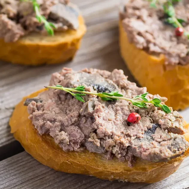 Slices of bread topped with beef liver pate, garnished with herbs and spices, placed next to a mushroom on a wooden surface.