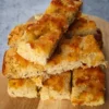 A stack of keto focaccia slices on a wooden board with melted cheese and herbs.