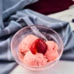 A clear glass bowl with three scoops of creamy keto strawberry crème fraîche ice cream, garnished with a fresh strawberry slice, placed on a white marble surface with a gray napkin in the background.