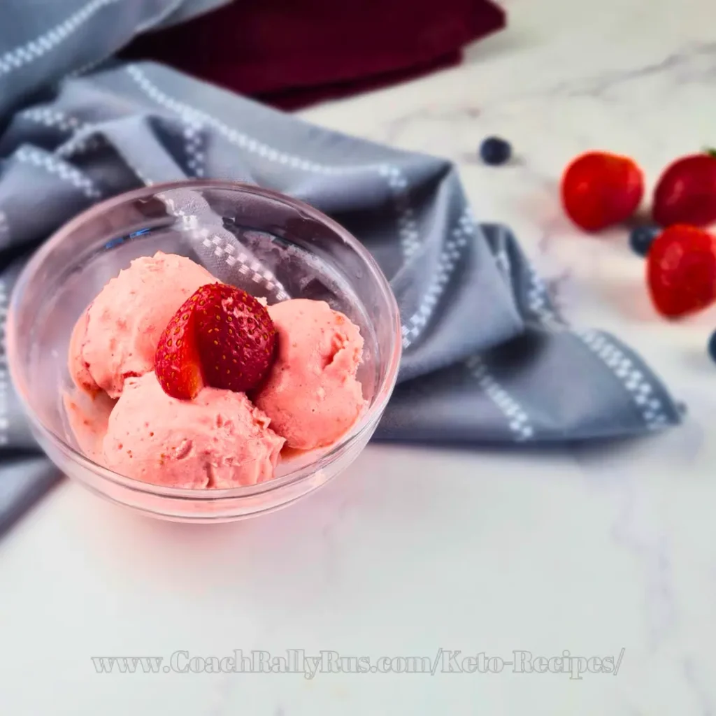 A clear glass bowl with three scoops of creamy keto strawberry crème fraîche ice cream, garnished with a fresh strawberry slice, placed on a white marble surface with a gray napkin in the background.
