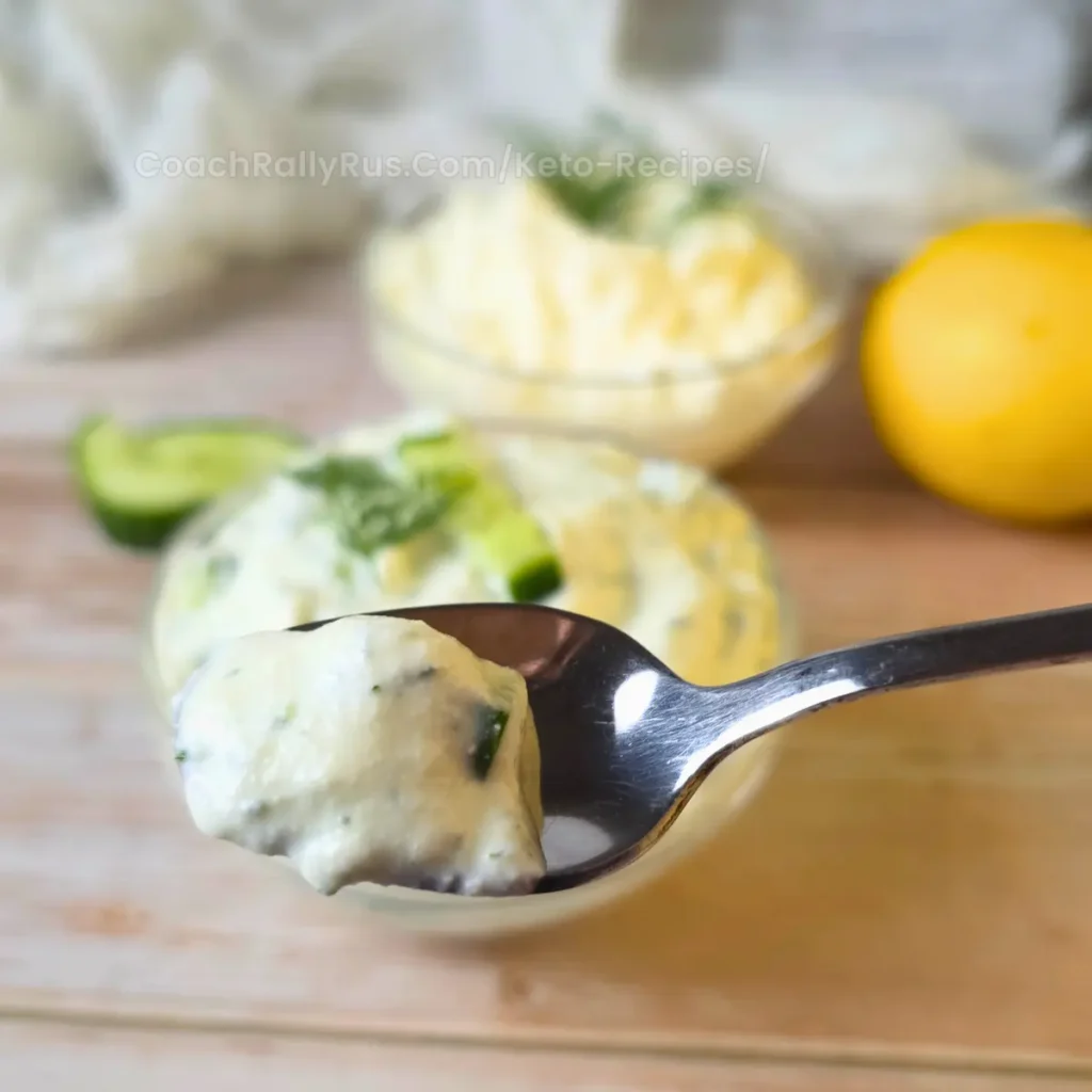 A close-up of a spoonful of creamy dill cucumber cottage cheese dip, with a bowl of dip, fresh cucumber, and lemon in the background.
