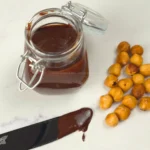 Keto Nutella in glass jar with roasted hazelnuts and chocolate spread on knife