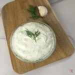 A close-up of creamy garlic dill yogurt sauce garnished with fresh dill in a small glass bowl on a wooden board