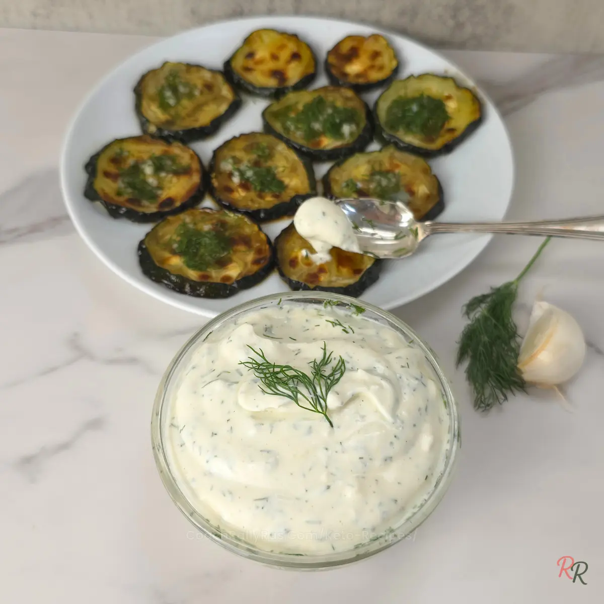 A bowl of garlic dill yogurt dip next to roasted zucchini rounds on a plate, with a spoonful of sauce being served
