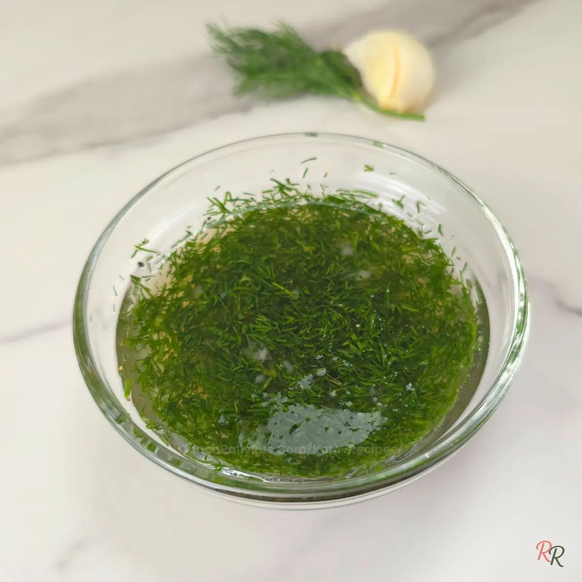 Overhead shot of garlic dill vinegar sauce in a clear bowl with fresh dill and garlic in the background