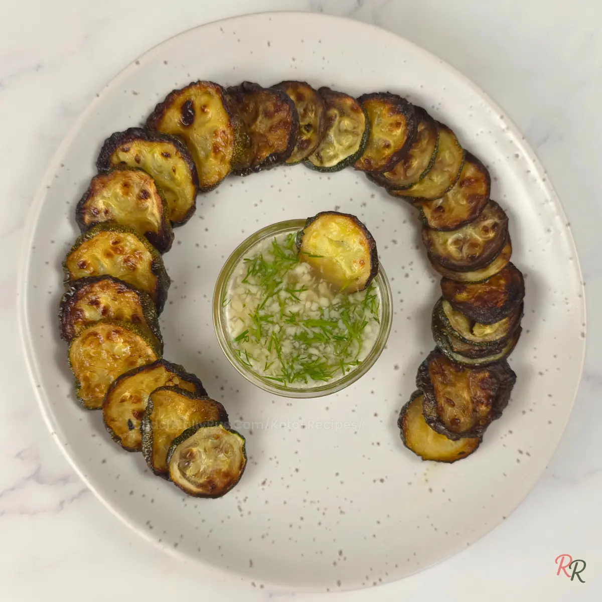 Spiral arrangement of crispy zucchini rounds on a plate with a bowl of garlic dill sauce in the center.