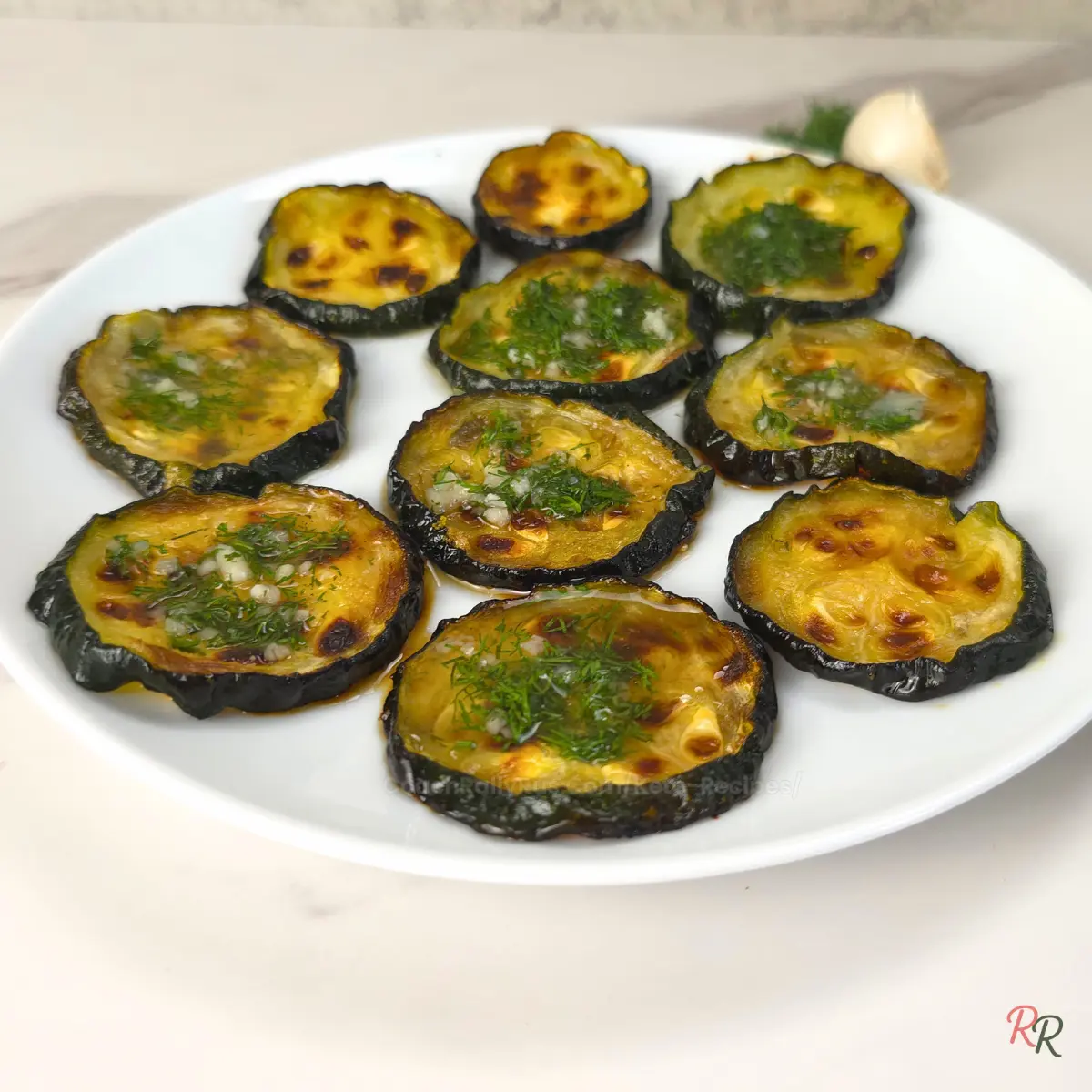 Side angle of golden brown zucchini slices with fresh dill and garlic on a white plate.
