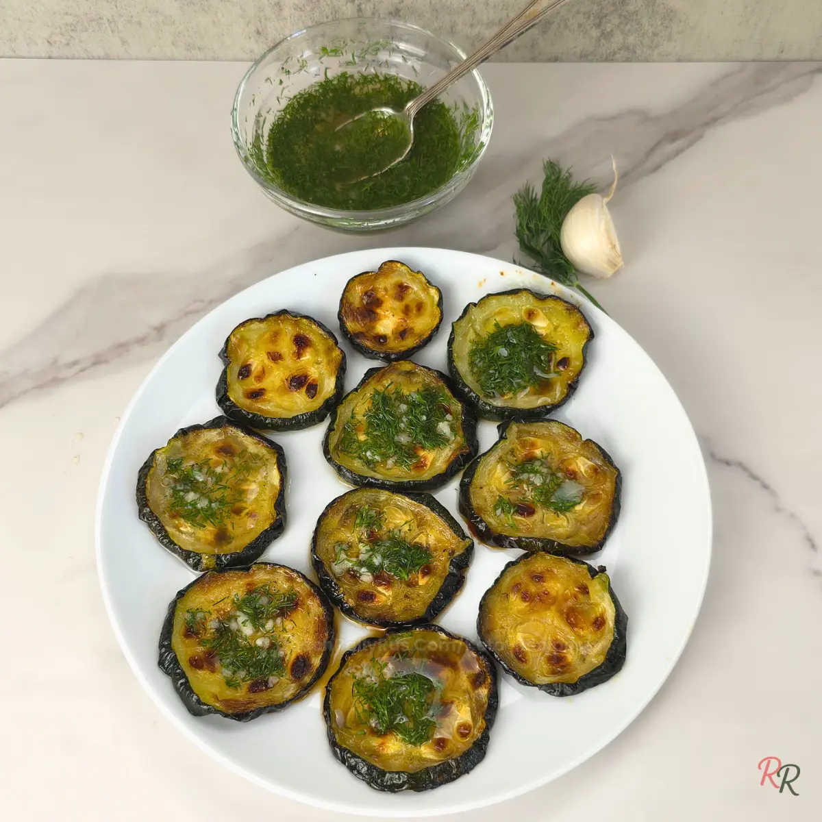 Flatlay of garlic dill roasted zucchini served on a white plate with extra sauce in a bowl.