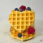 Heart-shaped waffle topped with berries in front of stacked waffles, with scattered blueberries