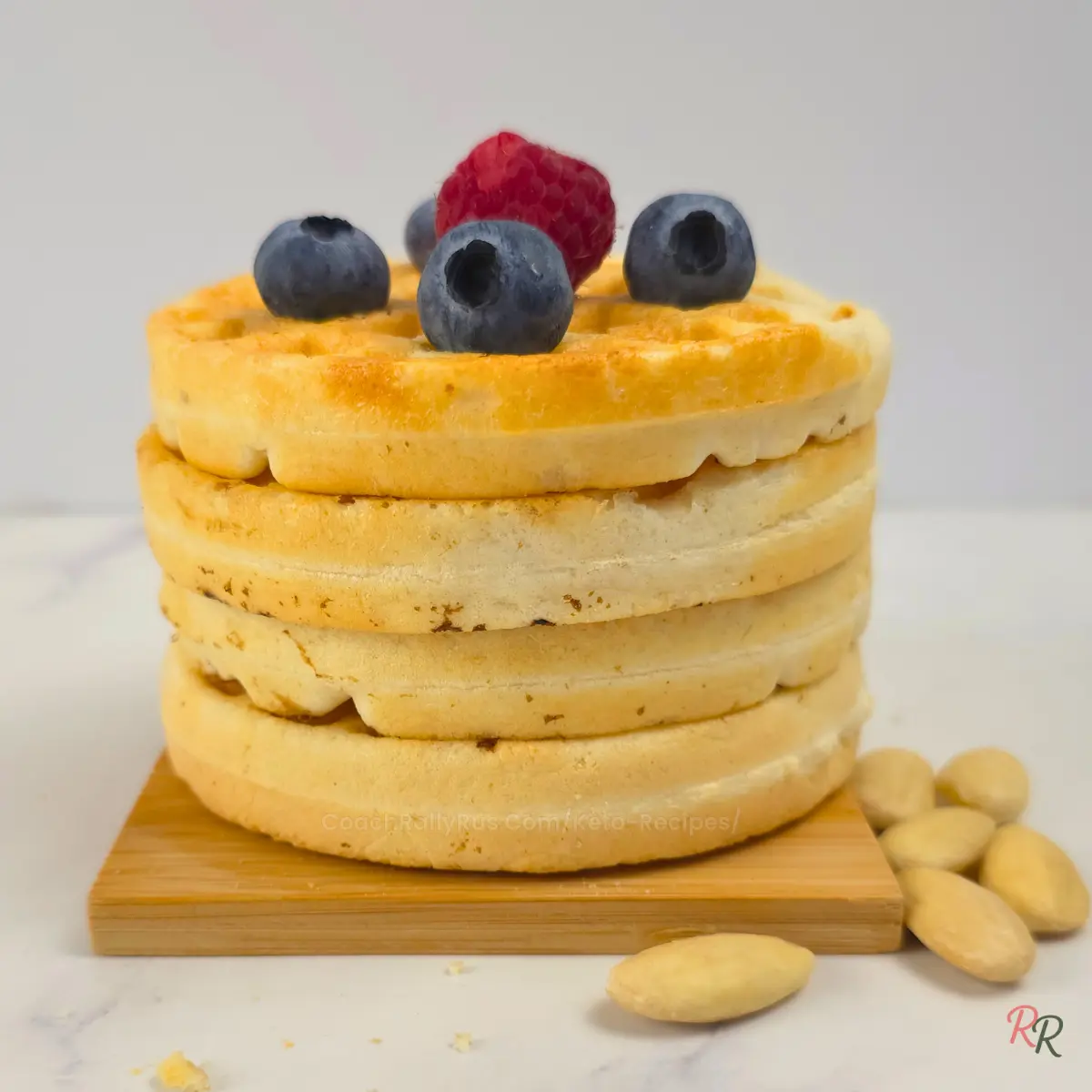 Tall stack of fluffy almond flour waffles topped with fresh blueberries and raspberry, with almonds on the side.