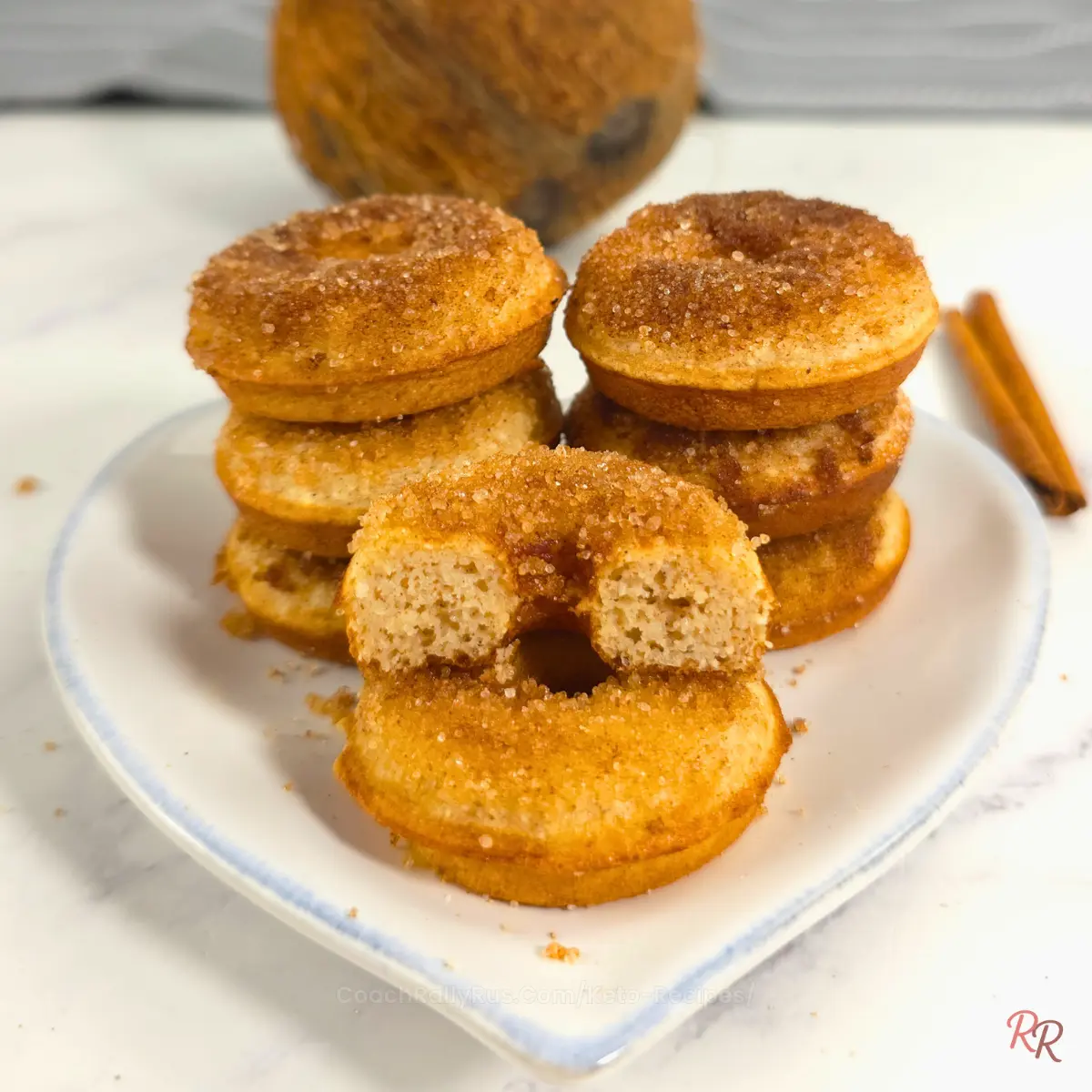 Close-up of keto cinnamon sugar donuts stacked on a plate with a coconut and cinnamon sticks in the background.
