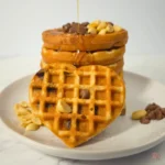 Heart-shaped keto cookie waffle topped with chocolate chips and peanuts in front of a waffle stack by Rally Rus
