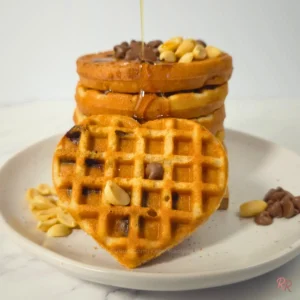 Heart-shaped keto cookie waffle topped with chocolate chips and peanuts in front of a waffle stack by Rally Rus