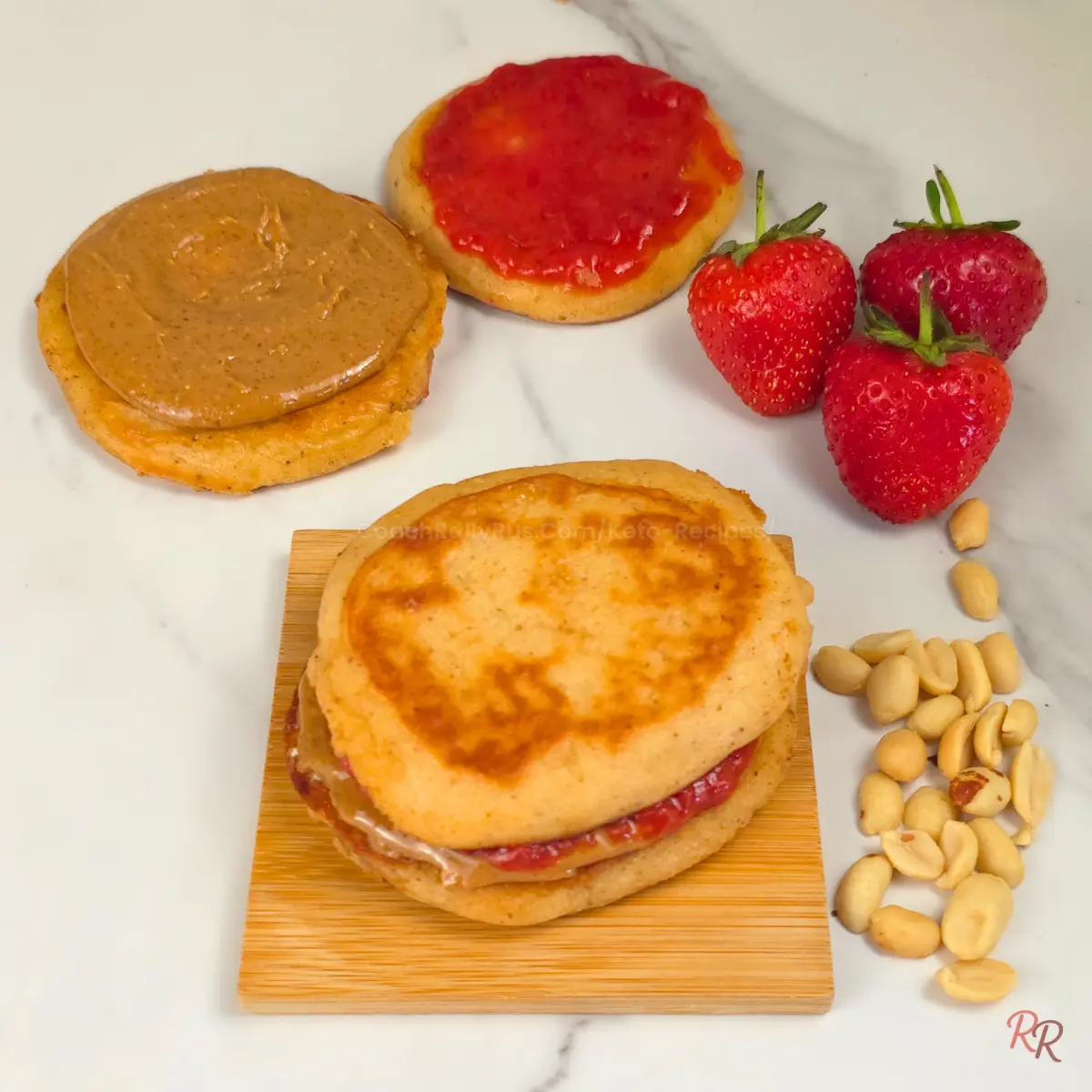 Assembly of keto pancake sandwiches with peanut butter and jam, shown open-faced next to fresh strawberries and peanuts.