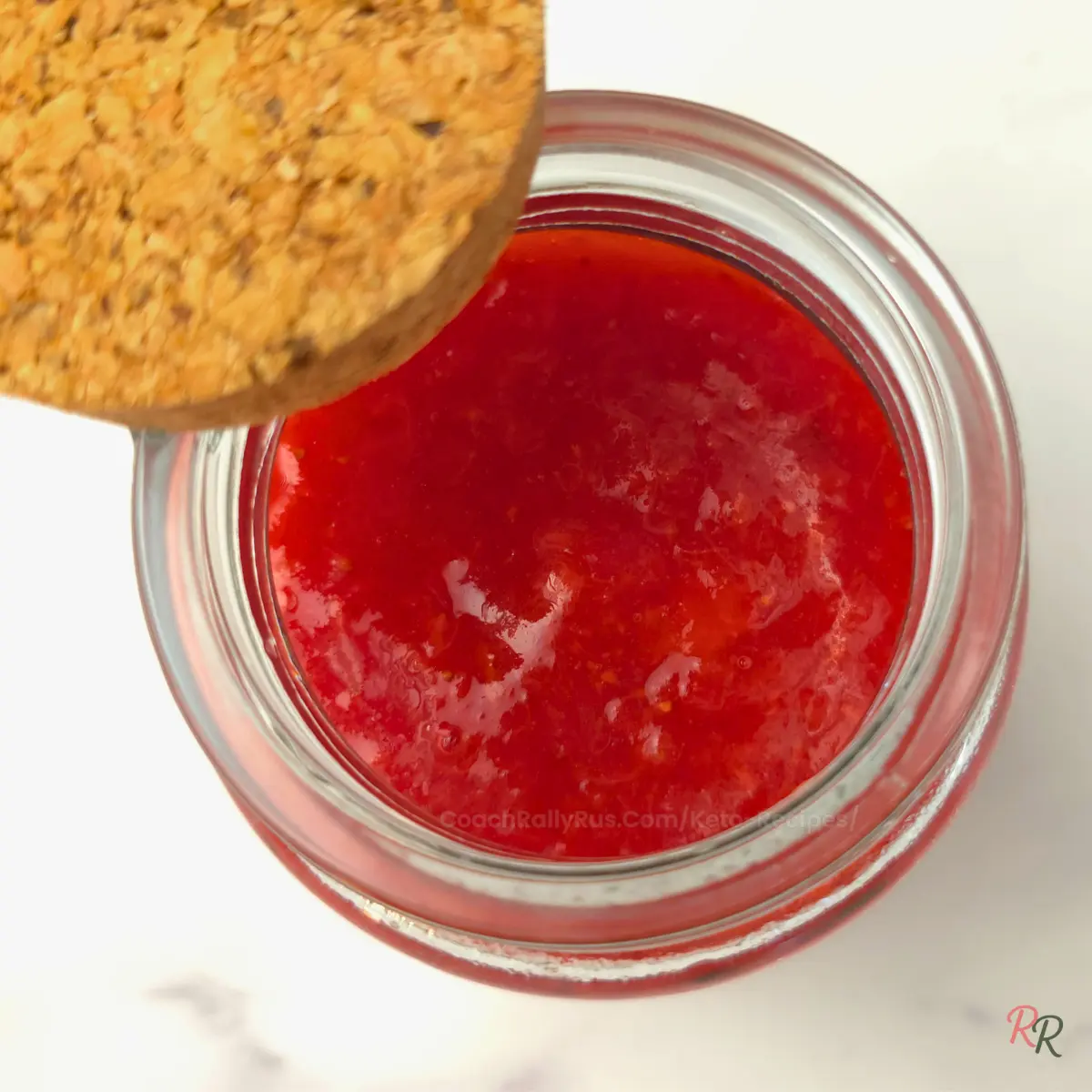 Glass jar of keto strawberry jam with cork lid next to whole strawberries and lemon.