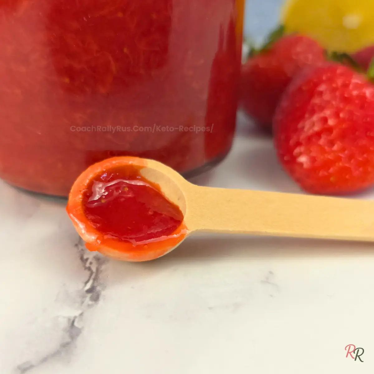 Close-up of keto strawberry jam on a spoon in front of a jar with strawberries and lemon.