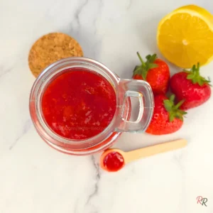 Overhead shot of homemade keto strawberry jam in a glass jar with lemon and strawberries on the side.
