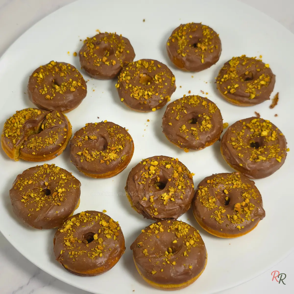 batch of keto pistachio almond flour donuts on a white plate, glazed and topped with chopped pistachios.