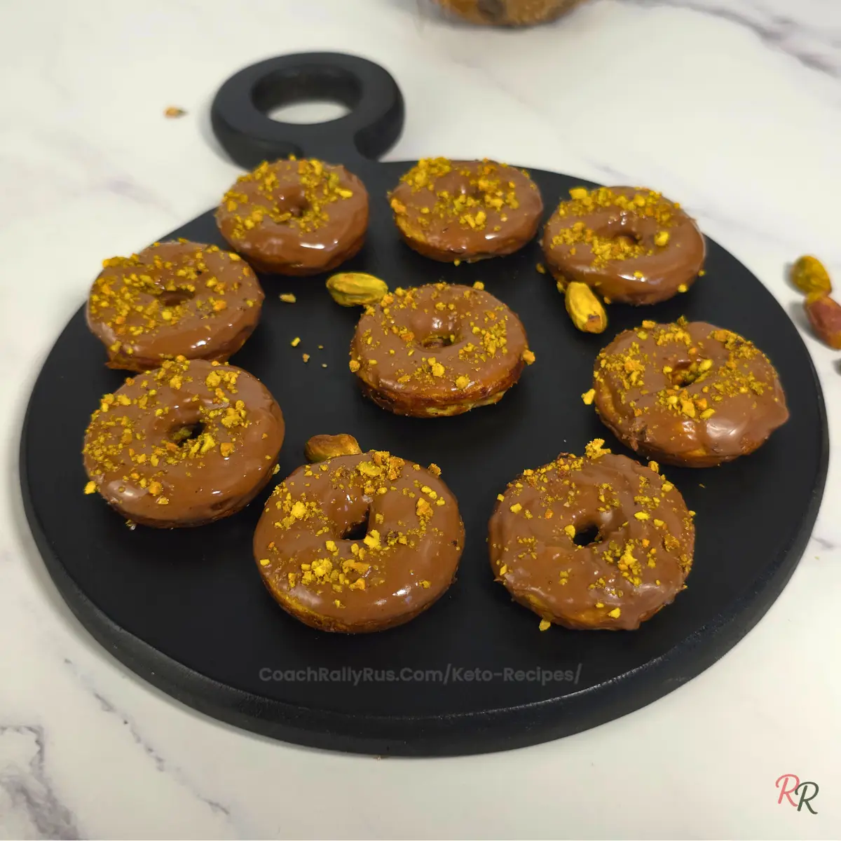 Nine keto pistachio donuts with a golden brown finish and chocolate glaze arranged neatly on a round black tray.