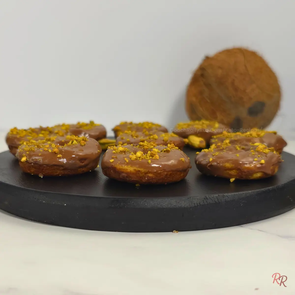 Top-down view of nine keto pistachio donuts arranged on a round black tray with scattered pistachios on the side.