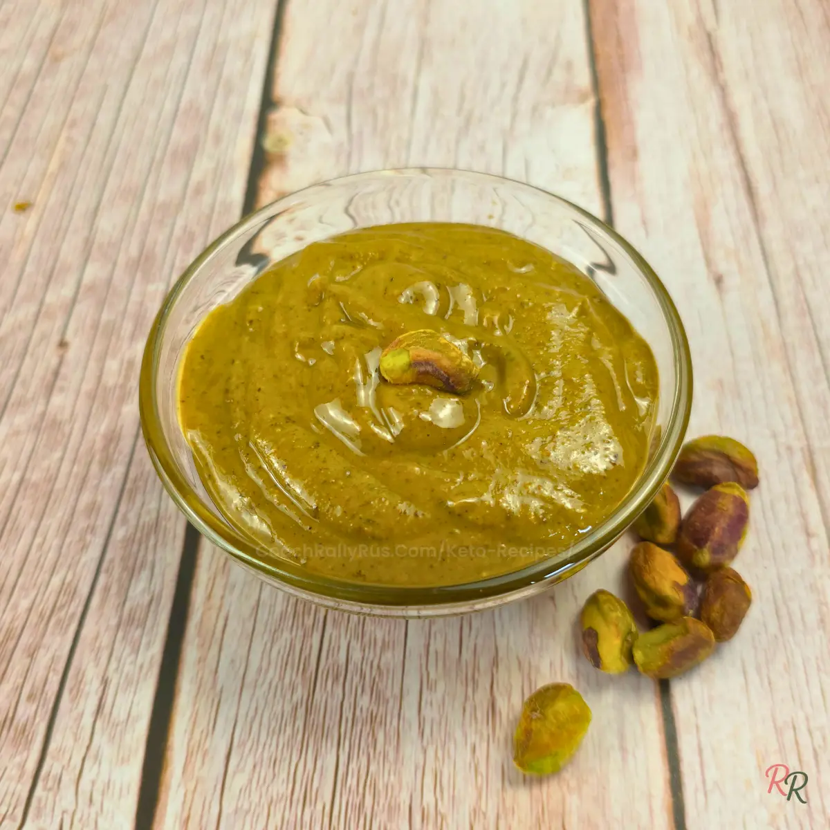 Small glass bowl of pistachio butter on a rustic wooden background, with scattered pistachios beside the bowl