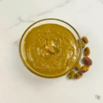 Glass bowl of sugar-free pistachio butter on a white marble background, topped with a single roasted pistachio and surrounded by whole nuts